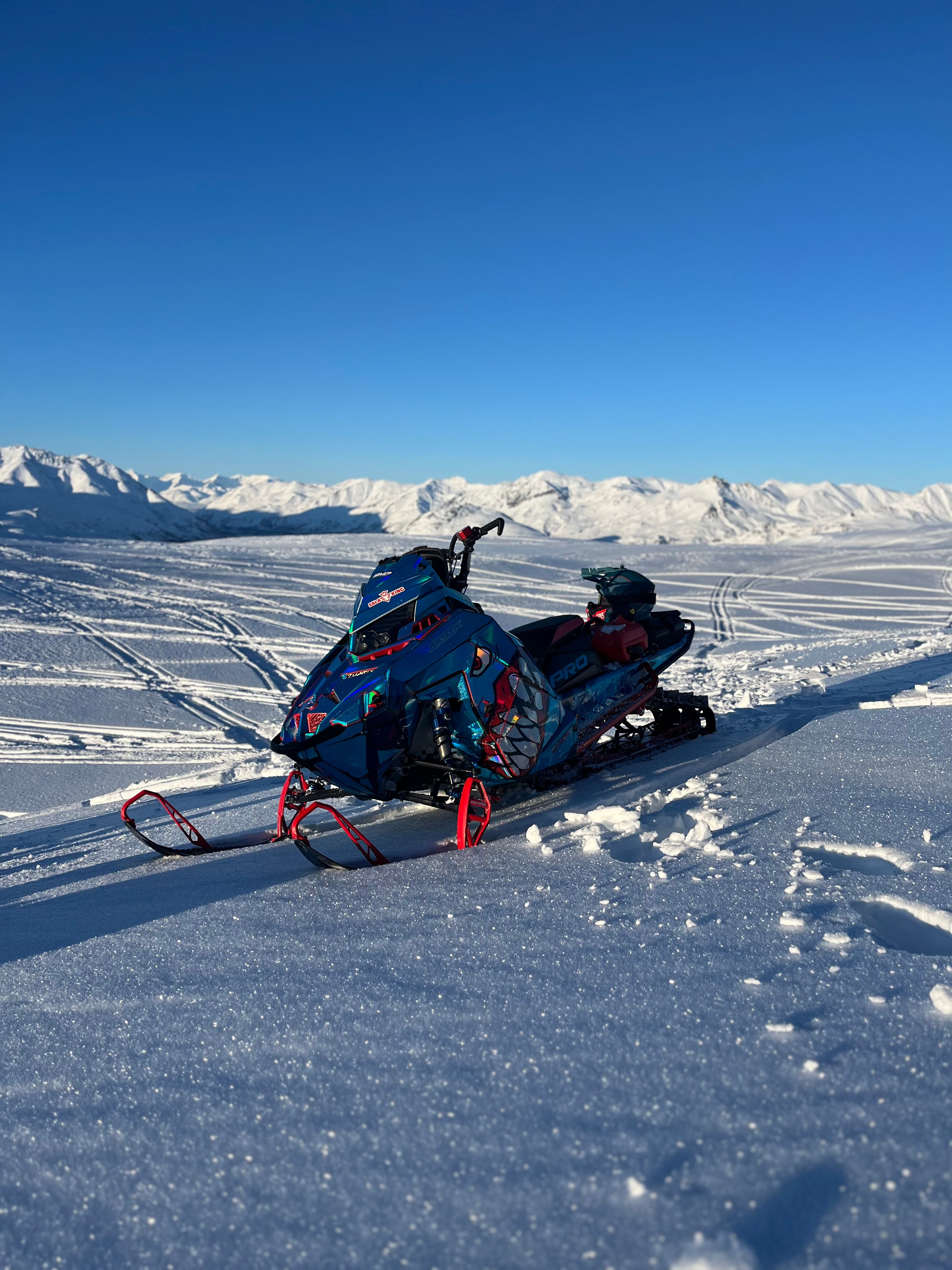 Blue snowmobile with a Prismatic Chrome sled wrap  on a snowy landscape with mountains in the background