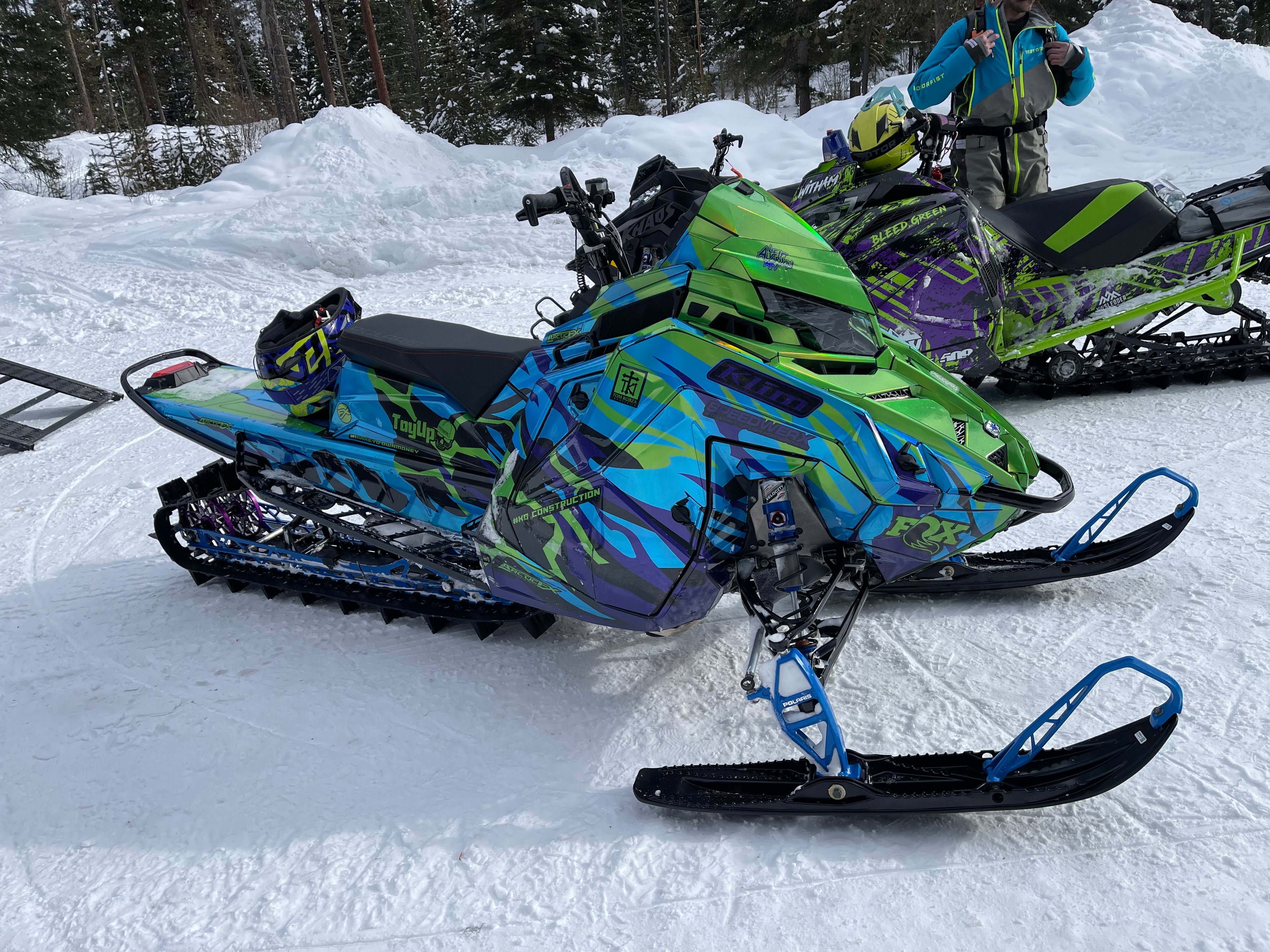 Colorful snowmobiles parked on a snowy surface with a person in the background.