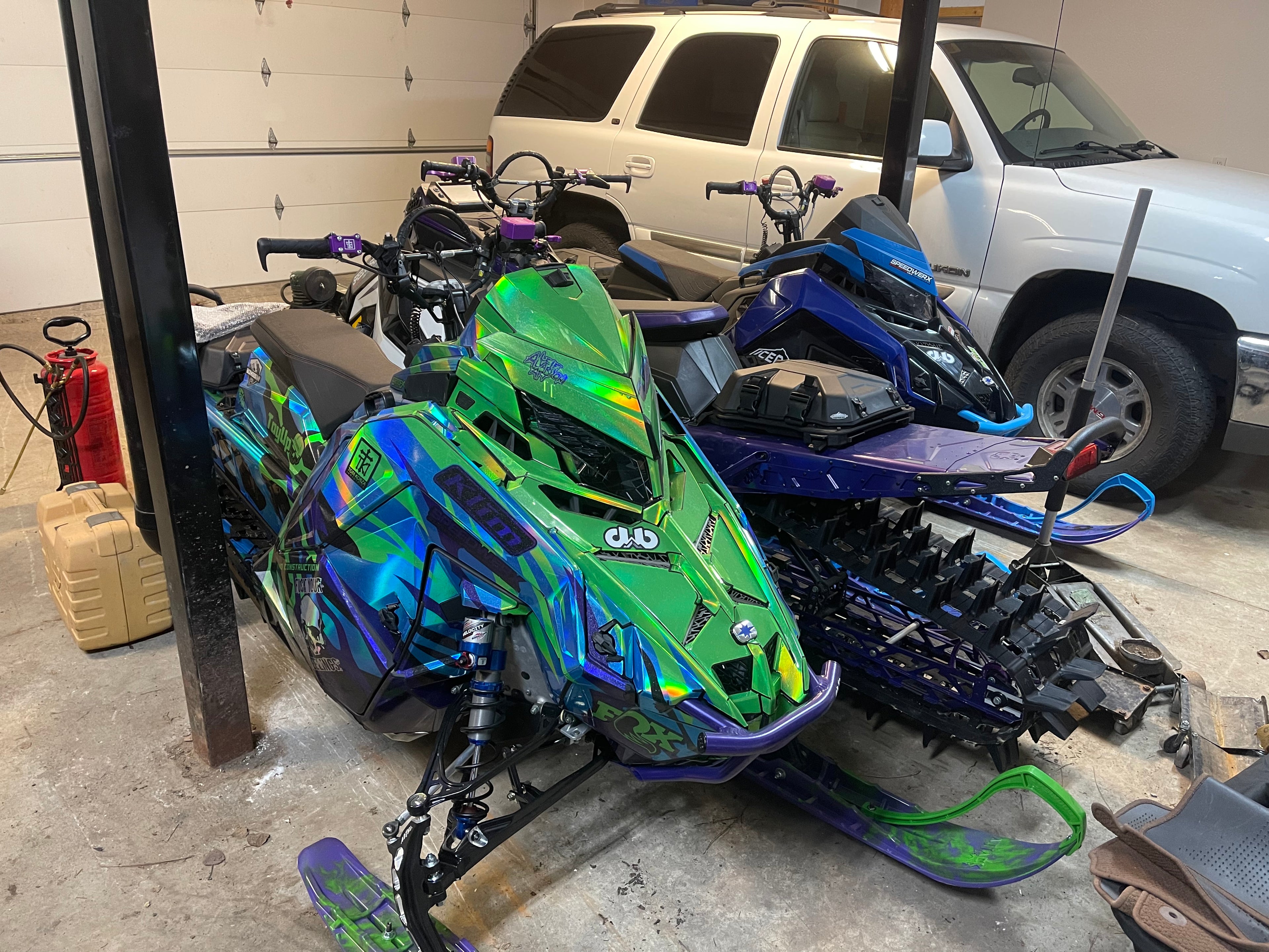 Colorful snowmobiles in a garage with a white SUV in the background.
