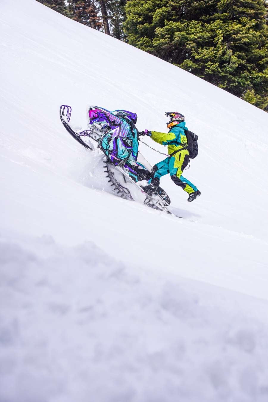 A snowmobile with a rider in a bright turquoise and yellow snowsuit navigating deep snow on a snowy mountain slope, with evergreen trees in the background.